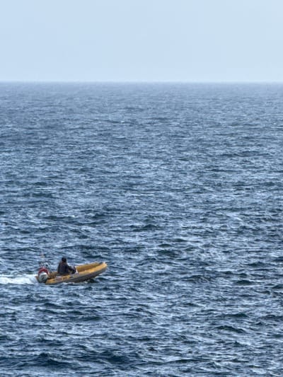 A person is pictured in a kayak on a massive waterfall. The kayak is yellow with a black seat and a person wearing dark clothing and a helmet. The waterfall is immense, appearing to cascade endlessly, with a textured, blue-gray surface. To the left of the waterfall is a vast expanse of pale, hazy sky, suggesting an overcast or foggy day. The image captures a moment of extreme sport or adventure, highlighting the sheer scale of the natural phenomenon and the smallness of the human participant. The orientation of the image suggests it was taken from above or at an angle looking down the waterfall.