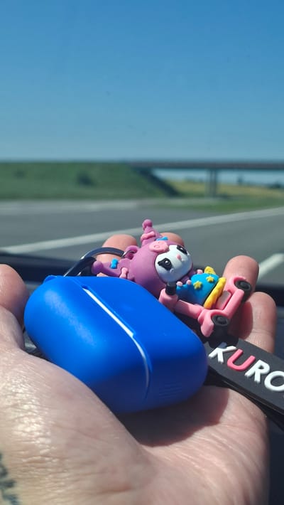 A close-up shot features a person's hand holding a blue object, possibly an earbud case, and a pink keychain figurine of a character with large eyes and a unicorn horn. The figurine is positioned on a small pink skateboard with yellow accents and blue wheels. A black tag with white and pink lettering, appearing to say "KURO," is attached to the keychain. In the background, a highway stretches into the distance under a clear blue sky, with a green embankment and a bridge visible. The overall impression is a roadside scene, potentially in or near Станькаўскі сельскі Савет, Belarus, given the context of the location.