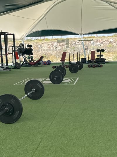 The scene depicts an outdoor fitness facility or gym located in Las Crucitas, Mexico. The ground is covered with green artificial turf, featuring white markings, possibly a logo or boundary lines. The area is covered by a large, white fabric tension structure or canopy, providing shade.
In the foreground, two barbells are positioned horizontally on the turf. The closest barbell has black weight plates, with "10 KG" visible on one plate. A second barbell, further back, is also loaded with black weight plates. Extending into the midground, additional barbells with black plates are arranged in a line.
To the left, a black metal squat rack or power cage is partially visible. Further back, a red-seated leg press machine is present, along with several red and blue medicine balls. In the center-right, two red adjustable weight benches are visible, one positioned near a rack containing multiple red dumbbells. Several tall, black vertical poles, consistent with sled push handles or similar equipment, are also present.
The background reveals a natural environment beyond a clear barrier, likely glass or transparent fencing. This barrier separates the gym from an undeveloped hillside or embankment composed of dry earth and sparse vegetation, indicating an arid or semi-arid climate. The sky above is bright and clear. No individuals are present in the image.