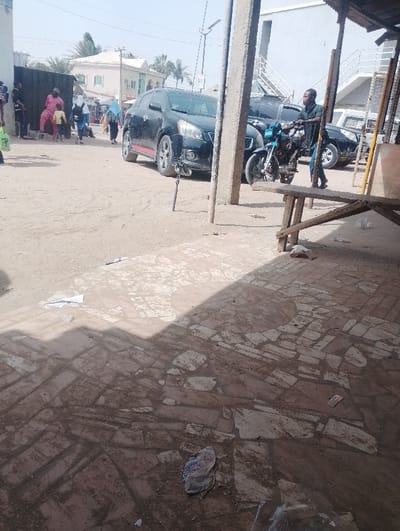 This image presents an outdoor urban scene in Panshekara, Nigeria, under bright daylight. The ground consists of compacted dirt mixed with irregular, broken paving stones, exhibiting scattered litter including plastic bags and paper. A distinct, diagonal shadow line from an unseen overhead structure bisects the mid-ground.
In the mid-ground, a black SUV with red lower body accents is parked, facing left. Behind it, a second dark-colored SUV is partially visible, followed by a lighter-colored vehicle further back. A male subject, dressed in a dark short-sleeved shirt and blue trousers, is actively pushing a light blue motorcycle with a black seat, moving from right to left. To the far right, a robust wooden or concrete pole stands vertically, partially obscuring two roughly constructed wooden structures, possibly benches or tables, in the foreground.
On the left side of the frame, a group of approximately 5-7 individuals is gathered near a low, dark wall or fence. This group includes a person in a prominent pink/red outfit accompanied by a child in similar attire, and another person wearing a light blue head covering over dark clothing, carrying a backpack. In the background, a light-colored building with a dark roof and palm trees are visible on the left, while a multi-story building with exterior stairs is present on the right. A street light pole with an attached luminaire rises in the central background.