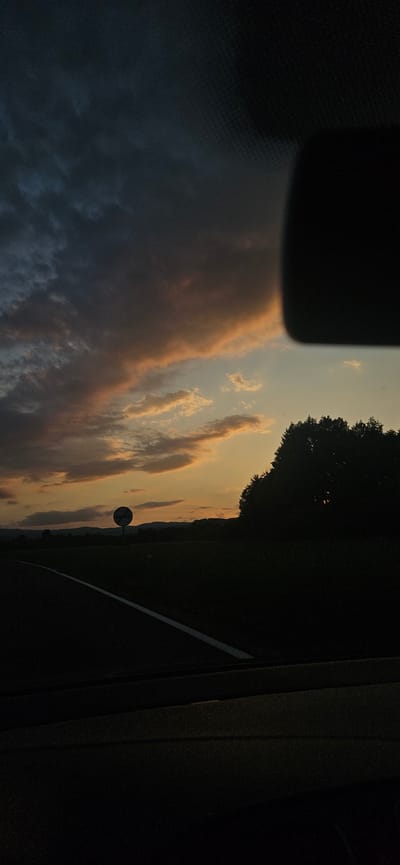The image captures a sunset landscape as viewed from inside a moving vehicle, located in the city of Dolna banya, Bulgaria. The upper portion of the frame is dominated by a twilight sky, transitioning from dark blue-grey, heavy clouds in the upper left to vibrant orange, yellow, and peach hues among lighter clouds towards the horizon on the right, indicating a setting sun.
In the immediate foreground, the dark silhouette of a vehicle's dashboard or interior is visible along the bottom edge. A white solid line marks the edge of a paved road, which curves gently towards the left in the lower-left quadrant. To the right of the road, the landscape consists of dark, silhouetted fields and a dense line of trees forming a dark mass against the illuminated sky in the mid-right. A distinct, round road sign with a pole is visible in the mid-ground, centered horizontally, and appears largely silhouetted, obscuring its specific symbol. Distant hills or mountains form a faint, dark silhouette against the horizon beneath the sunset clouds. A dark, rectangular object, possibly a rearview mirror or part of the car's interior roof lining, intrudes into the upper-right corner of the frame. No individuals are present in the scene, and no specific actions or interactions are depicted.