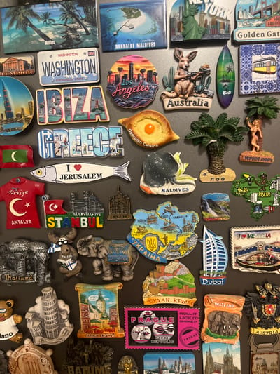 The image is a close-up, overhead shot of a collection of refrigerator magnets displayed on a dark, flat surface, likely a refrigerator door. The magnets are varied in shape, size, and subject matter, depicting landmarks, flags, and cultural motifs from numerous international locations. Several magnets feature text in various languages, indicating origins such as "Maldives," "Washington," "Ibiza," "Greece," "Australia," "Bali," "Lisboa - Portugal," "Jerusalem," "Türkiye," "Antalya," "Istanbul," "Milano," "Maldives," "Brazil," "Ukraine," "Dubai," "Venice," "Finland," "Sri Lanka," "Thailand," "Czech Republic," and "Austria." Other magnets depict stylized interpretations of famous structures like the Eiffel Tower (though not explicitly labeled), the Leaning Tower of Pisa, and the Burj Al Arab. There are also magnets with animals, including elephants, a kangaroo, a moose, and a fish. The overall impression is a personal collection of travel souvenirs. The location of this collection is not identifiable from the image content itself; there are no visual cues to suggest it is in Novaya Adygeya, Russia.