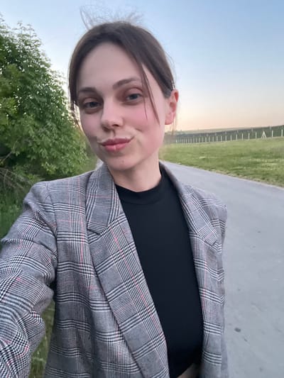 A young woman is posing for a selfie outdoors. She has light skin, dark hair pulled back from her face, and is wearing a black top and a grey plaid blazer. The background includes a concrete path, green grass, trees, and a fence in the distance. The sky is a pale blue. The location appears to be a park or recreational area. The image provides insufficient data to concretely deduce its location in Indjija Municipality, Serbia.