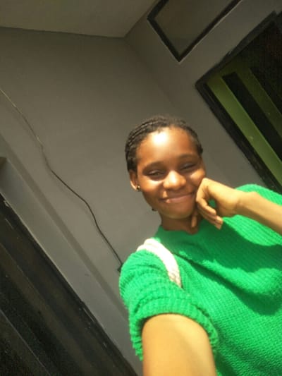 A young Black female is depicted in a close-up, upward-angled portrait. She is situated in an indoor or semi-sheltered environment, the specific urban location of Cotonou, Benin, not visually verifiable from the image content. The subject, appearing to be in her late teens or early twenties, smiles broadly with slightly squinted eyes and her chin resting lightly on the knuckles of her left hand. Her dark hair is styled in tight, short braids or cornrows. She wears a vibrant green, short-sleeved, ribbed or knitted top, with a thick white strap, likely from a bag, visible over her right shoulder. A small dark stud earring is present in her left ear. The background features a light gray wall, with a dark, angular opening or frame visible in the upper right quadrant. A thin, dark cable runs diagonally across the upper left section of the wall. The scene is illuminated by bright, direct light, casting soft shadows.