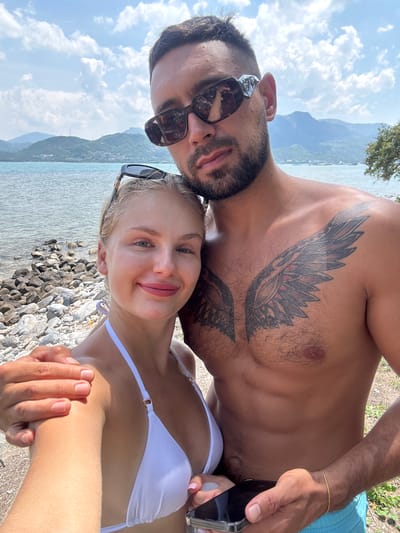 The image shows a selfie taken by a man and a woman on a beach. The man is shirtless, revealing a large tattoo of wings on his chest. He is wearing sunglasses and blue swim trunks. The woman is wearing a white bikini and sunglasses. They are standing close together, with the man's arm around the woman's shoulder. In the background, there is a body of water, a rocky shore, and mountains. The sky is blue with some clouds. The location is identified as Ninh Ích, Vietnam.