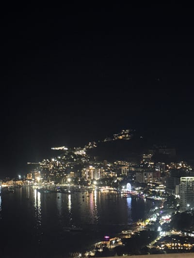 This is a nighttime aerial photograph of a coastal city with a bay filled with boats. The city is built on a hillside, with buildings illuminated by lights that create a warm glow against the dark sky. A Ferris wheel is visible in the distance, adding a festive element to the scene. The lights from the city reflect on the water, creating a shimmering effect. The image captures a vibrant and lively urban environment at night, with the natural beauty of the coastline. The location of Košljun, Montenegro is suggested in the prompt but not definitively identifiable from the visual information alone. The scene is composed of buildings, a body of water, boats, and the night sky. There are no visible people or specific actions.