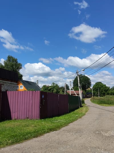 The image depicts a rural scene on a sunny day in Venyukovo, Russia. A light gray gravel road curves to the right, bordered by a lush green grassy area on the left. A tall, reddish-purple corrugated metal fence runs along the left side of the road, with a section of green fence further down. Beyond the fence, there is a residential property with a light-colored brick building with a dark metal roof and an orange wooden house with a dark green roof. Trees and other foliage are visible in the background. Power lines and utility poles with streetlights are also present, stretching across the sky. The sky is a vibrant blue with scattered white cumulus clouds. The overall impression is of a quiet, well-maintained rural settlement.
