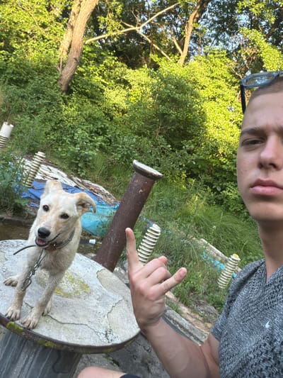 The image shows a young person taking a selfie outdoors with a dog. The person is wearing glasses and a grey t-shirt, and they are making a hand gesture. The dog is light-colored and appears to be a mixed breed, wearing a collar and leash. The dog is standing on a round, stone-like surface, possibly a table, next to a dark brown, cylindrical object.
The background is lush and green, with dense foliage from trees and grass. Some white, coiled objects are visible in the background, which could be part of some garden decoration or discarded items. The scene suggests a casual, outdoor setting, perhaps a garden or backyard, on a sunny day given the lighting. The overall mood appears relaxed and playful, with the person smiling towards the camera and the dog looking alert. The image has a slightly tilted orientation. The location context is Dolno Sahrane, Bulgaria. There is no visible text in the image.