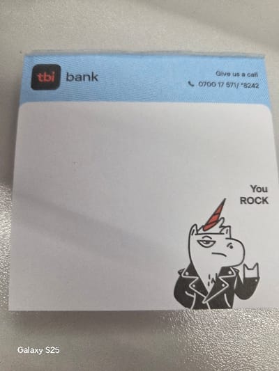 A square-shaped TBI Bank sticky note pad is depicted resting on a light-colored, textured surface within the city of Novo Delchevo, Bulgaria. The top section of the pad features a light blue band. On the upper left, the "tbi bank" logo is present, with "tbi" in a black square on a red background, followed by "bank" in black text. On the upper right, the phrase "Give us a call" is printed above a telephone icon and the contact number "0700 17 571 / *8242". The main body of the pad is white. In the lower right quadrant, a cartoon illustration of a white unicorn with a red horn and a black leather jacket is shown making a "rock-and-roll" hand gesture. To the left of the unicorn, the words "You ROCK" are printed vertically in black capital letters. The text "Galaxy S25" is visible in the bottom left corner of the image, indicating the device used to capture the photograph.