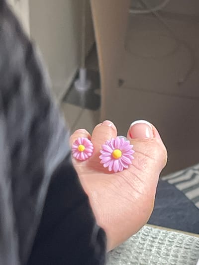 This image depicts a close-up view of a person's toes adorned with two decorative flower-shaped charms. The charms are pink with yellow centers and appear to be made of plastic or resin. One charm is positioned on the big toe, and the other is on the toe next to it. The toenails have a French manicure. The background is blurred, showing a dark fabric on the left and a light-colored surface with a striped pattern on the right. The scene is likely indoors. There is no discernible information in the image to indicate a location in Montrond-les-Bains, France.