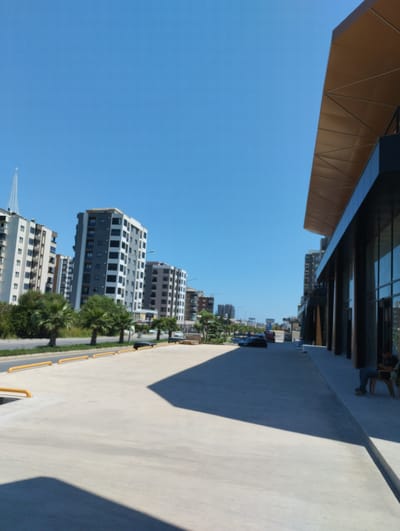 The image depicts an outdoor scene in Toroslar, Turkey, on a clear, sunny day. In the foreground, a vast, light-colored concrete expanse, possibly a parking lot or plaza, dominates the lower portion of the frame, with yellow bollards lining the edge. To the right, a modern building with dark columns and a large glass facade is visible, featuring a striking geometric canopy overhead. A person is seated on a chair on the sidewalk in front of this building.

Further back, a street runs horizontally across the midground, lined with lush green trees and palm trees. Beyond the street, a row of multi-story apartment buildings stands, varying in architectural style and color, with many featuring balconies and gray and white facades. The sky above is a brilliant, cloudless blue. In the distance, more buildings and structures are visible, suggesting a developed urban environment. The overall impression is one of modern construction and urban living.