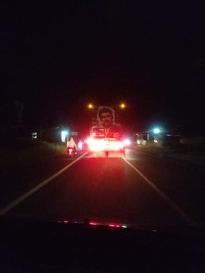 This is a night-time, eye-level, vertical, point-of-view photograph taken from inside a vehicle. The scene depicts a road at night, likely in Meunasahpulo, Indonesia, given the location context. The road is illuminated by the headlights of oncoming vehicles and streetlights. In the center of the image, a large billboard featuring a man's face is prominently displayed and lit up. The man has a mustache and is wearing a patterned shirt.
In front of the billboard, what appears to be a truck or large vehicle is visible, its bright headlights creating a strong glare that obscures some details of the road ahead. To the left of the road, there is a person riding a motorcycle, also illuminated by headlights and taillights. The person is wearing light-colored clothing. The road has white lane markings.
The overall atmosphere is dark, with limited visibility beyond the illuminated areas. The bright lights create reflections on what may be a wet road surface or the vehicle's windshield, adding to the blurred and atmospheric quality of the image. The time of day is clearly night, and there are no obvious indicators of specific weather conditions, though the reflections might suggest dampness. The scene conveys a sense of movement and travel through a dimly lit urban or semi-urban environment. There is no visible text in the image.