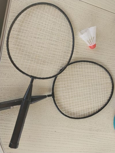 This image depicts a pair of black badminton rackets and a shuttlecock arranged on a light-colored wooden surface. The rackets are positioned diagonally, with one larger racket at the top and a smaller one below. The shuttlecock, with its white feathers and red base, rests to the right of the upper racket. The wooden surface exhibits a wood-grain pattern with visible lines and texture. The lighting suggests an indoor setting, possibly a table or floor. The image does not contain any information or context relating to the city of Nizhneye Myachkovo, Russia.