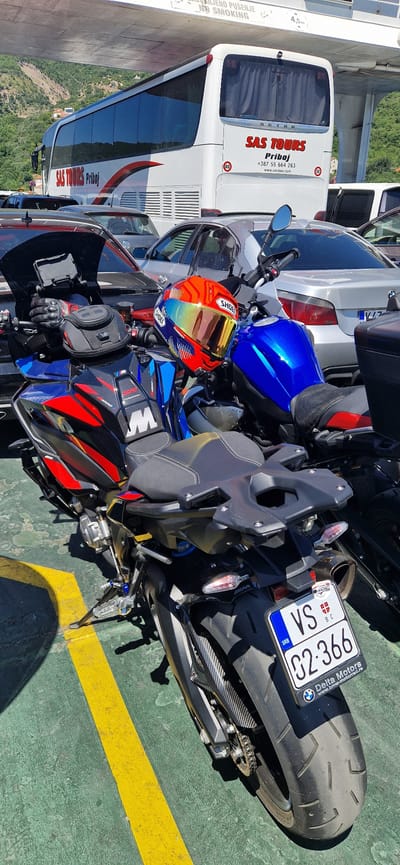 This media file captures a motorcycle parked on what appears to be a ferry deck, indicated by the green surface with yellow painted lines. The motorcycle is a prominent subject, featuring a blue fuel tank, a black seat, and a license plate with Serbian markings (SRB, VS, 02-366). A colorful helmet, possibly a Shoei, rests on the motorcycle's handlebars, showcasing a vibrant orange and blue design.
In the background, a white tour bus with "SAS TOURS Priboj" printed on its side is visible. The bus also has "SETRA" branding. Other vehicles, including a silver car with a BMW logo on its license plate frame, are partially visible, suggesting a transportation hub or a ferry carrying vehicles. The text "NO SMOKING" and "ANJENO PUŠENJE" is present on a white vertical element on the left side of the frame, along with a height indicator of "4.5m". The surrounding environment suggests a sunny day with hints of greenery and distant hills in the background, possibly indicating a coastal or mountainous region. The overall scene depicts a moment of transit, with vehicles waiting to embark or disembark.