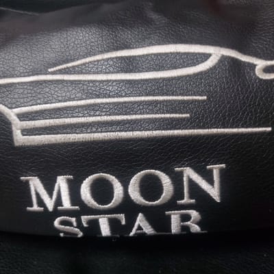 A close-up image reveals a black, finely textured surface, consistent with faux leather or leather material, located in Gimpo City, South Korea. Embroidered in white thread are two distinct elements. The upper element is a stylized, elongated silhouette of a car, depicted in profile and facing right, with horizontal lines forming its body. Below this emblem, the words 'MOON STAR' are embroidered in two lines of capital letters, with 'MOON' positioned above 'STAR'. No individuals, interactions, or specific environmental details beyond the textured material are discernible.