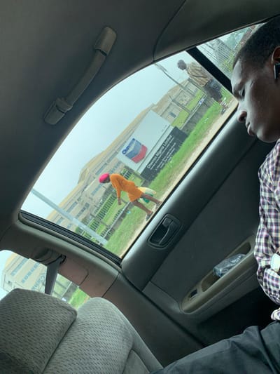 The image, taken from inside a vehicle, shows a young man in the foreground, possibly sleeping, with part of his face visible. Through the sunroof and side window, a Chevron Nigeria Limited building and a sign with the Chevron logo are visible. A woman in an orange dress and pink headscarf is walking in front of the building. Another person, wearing a striped shirt, is standing near a fence in the background. The environment appears to be an industrial or commercial area with greenery and a cloudy sky. The vehicle's interior, including seats and a handle, is also visible. The location is identified as Ojomu, Nigeria.