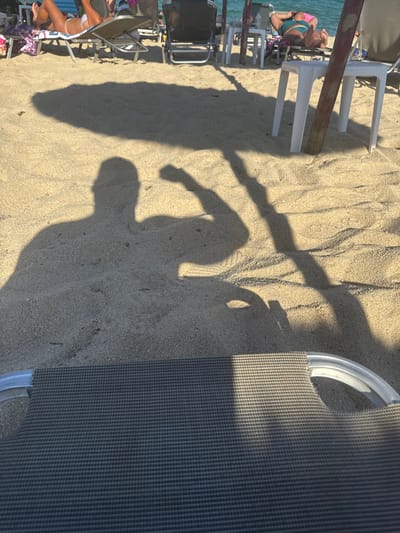 In Toroni Municipal Unit, Greece, on a sunny beach, a person's shadow is cast on the sand, flexing their bicep. The shadow is large and prominent, indicating a strong light source, likely the sun, from behind the subject. In the background, other beachgoers relax on lounge chairs. The scene is set with the gentle waves of the sea visible in the distance, hinting at a serene and leisurely atmosphere.