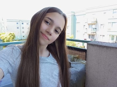 A young girl with long, brown hair, wearing a light grey t-shirt, is taking a selfie on a balcony. She is positioned in the center of the frame, with her face tilted slightly upwards and to her left. Her lips are slightly parted in a gentle smile, and she is wearing a small hoop earring in her left ear. Behind her, there are apartment buildings and trees. The balcony railing is visible in the foreground, and a textured concrete wall is on the right side of the image. The overall lighting suggests it is daytime. This scene is situated on a balcony in the city of Radzymin, Poland.