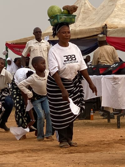 A woman wearing a Balenciaga t-shirt and black-and-white striped skirt carries a basket of fruit on her head. Several people are seated below, including a child in a white top and blue jeans. Others are dressed in lighter colors. The setting appears to be outdoors, possibly a market or event. A tent structure is visible in the background.