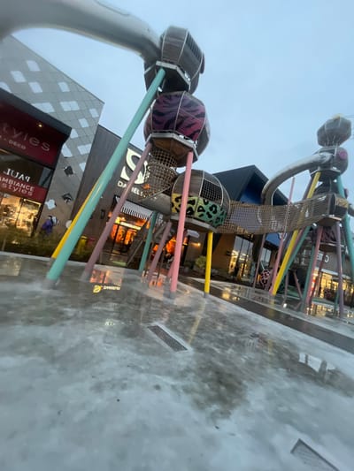 This image depicts an outdoor plaza area in Reichstett, France, under overcast, low-light conditions, likely twilight or early evening. The foreground features a light-colored, paved surface, which is wet and reflective, showing numerous reflections of artificial lighting from surrounding buildings. Several rectangular metal drainage grates are visible embedded in the pavement, and localized mist appears to be rising from the wet surface.

Dominating the midground is a large, multi-component playground structure or art installation. It consists of multiple pastel-colored support poles (light blue, pink, yellow, teal) that hold aloft various spherical or drum-shaped modules constructed from wooden slats and metal. Some modules are decorated with distinct animal print patterns, specifically pink and black zebra stripes, and green and black leopard spots. These modules are interconnected by mesh tunnels and bridges, and larger curved metal tubes or slides are also part of the configuration.

In the background, several commercial buildings are illuminated from within. To the left, a multi-story building features a patterned facade and large glass windows displaying storefronts with signs such as "Styles," "DECO," "PULP," and "AMBIANCE." One individual wearing a purple or dark blue jacket is partially visible near the "PULP" store, moving away from the camera. In the center, another building displays a prominent "S Daniel's" logo on its dark facade, with a red and white striped awning over a lower storefront labeled "DANIELL'S [STOP]". To the right, a third commercial building is partially visible with interior lighting and a partial sign reading "CINE...". Indistinct figures are visible within the brightly lit interiors of these background stores.