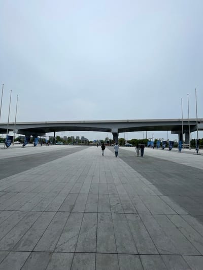 The image shows a large, empty plaza in Hangzhou, China, located at the coordinates 30.273 latitude and 120.417 longitude. The plaza is paved with gray concrete tiles and is bordered by a row of trees on both sides. A large, concrete bridge spans the plaza, creating a sense of depth and perspective. Several people can be seen walking on the plaza, with a few small cars parked on the sides. The sky is overcast, and the overall atmosphere is quiet and peaceful.