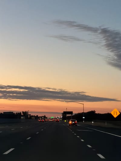 The image depicts a highway scene at dusk in Elk Grove, United States. The sky transitions from a pale blue at the top to orange and pink hues near the horizon, with wispy clouds present. A dark silhouette of trees lines the right side of the highway. On the road, several cars are visible, with their headlights and taillights illuminated. A yellow diamond-shaped sign with two vertical lines indicating lane reduction is posted on the right shoulder of the highway. Streetlights and traffic signals are also visible along the roadside. The perspective is from a moving vehicle, looking forward along the highway.