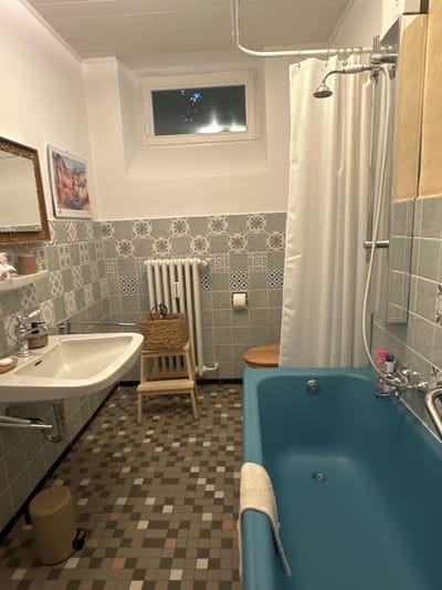 The image shows a small, tiled bathroom, likely in a residential setting, in Ammeldingen bei Neuerburg, Germany. The bathroom features a blue bathtub with a wooden toilet seat, a white sink with chrome fixtures, a white radiator, and a small stool with a woven basket. The walls are tiled with a patterned grey and blue tile design, and the floor is covered in a mosaic of small square tiles in shades of grey, pink, and white. A white shower curtain hangs from a metal rod. A small window with a slatted blind is visible at the top of the wall. The lighting appears to be artificial. There are no people or animals present in the image.