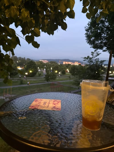 A close-up shot of a clear plastic cup with a brown liquid and a straw rests on a dark, textured glass table. The cup has a yellow logo on it. A small, square coaster is also on the table, featuring a floral pattern and the word "Koko" in Cyrillic, along with the text "для гостей кофеини". The background is an out-of-focus view of a park or outdoor seating area at dusk. Trees and pathways are visible, with scattered lights illuminating the scene. In the distance, buildings and hills are silhouetted against the twilight sky. The overall mood is calm and relaxed, suggesting an evening spent enjoying a drink outdoors. The time of day is likely early evening, as indicated by the dimming light and the park lights being on. The setting appears to be a cafe or restaurant with an outdoor seating area in Vladikavkaz, Russia, given the Cyrillic text and geographical context. No people are visible in the frame.