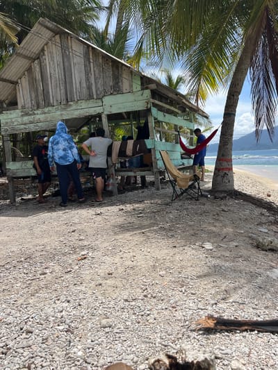 The image shows a group of people near a weathered wooden structure with a corrugated metal roof, possibly a small beach hut or shelter, in Babahie, Indonesia. The ground is covered with small stones and sand. Palm trees are visible, with one in the foreground to the left. In the background, the ocean meets the shore, and mountains are visible in the distance under a bright, cloudy sky.

Several individuals are gathered around the structure. One person, wearing a blue tie-dye hoodie and dark pants, stands with their back to the camera. Another person in a grey t-shirt and shorts stands nearby. Further into the structure, more people are visible, some appearing to be interacting or resting.

To the left of the structure, near the palm tree, a person in a blue shirt is lying in a red hammock. A beige camping chair is set up next to the hammock. The scene suggests a relaxed, tropical environment. The lighting indicates it is daytime, likely mid-morning or afternoon, with sunshine and shadows present. The presence of the beach, ocean, and palm trees clearly places the scene in a coastal, tropical setting. No visible text can be discerned.