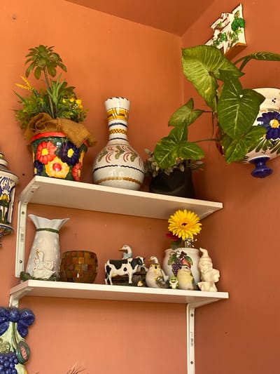 The image depicts a wall decorated with various ceramic figurines and vases, suggesting an indoor setting, likely a home or a shop in Resinera-voladilla, Spain. The subjects include several decorative items such as a large white vase with blue and yellow floral motifs, a smaller vase with green foliage and burlap accents, and a tall, slender vase with intricate patterns in yellow, blue, and brown.
A prominent feature is a white shelf unit on which several ceramic figurines are displayed. These include a gnome, a series of bird figurines in different poses, and a black and white cow figurine. Other decorative objects on the shelf include a small yellow daisy and a mosaic-like cylindrical object. At the bottom of the shelf, a white vase with duck figurines is visible, and further down, a decorative piece with grapes and other fruits can be seen.
The background is a textured, solid orange-brown wall. The lighting appears to be artificial, casting soft shadows and illuminating the objects clearly. The overall impression is one of a curated display of decorative and somewhat whimsical items. There is no indication of people or any specific activity taking place, making it a static scene focused on the objects. No visible text is present in the image.