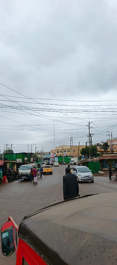 An eye-level view from a vehicle, likely a tuk-tuk, shows a bustling street scene under an overcast sky. A man in a black jacket stands in the road, with vehicles including a yellow and silver car nearby. Shops line the roadside, and people are engaged in various activities. Power lines crisscross the sky above buildings and signage. A Shell gas station sign is visible in the background, adding to the busy, urban environment.