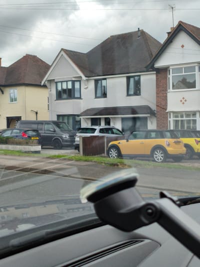 A daytime, medium-shot, exterior view shows a residential street scene in Wychavon, United Kingdom. The focus is on several parked cars, including a black van, a yellow Mini Cooper, and a silver SUV, positioned in front of residential properties with overcast skies above. The foreground of the image is dominated by the interior of a vehicle, with a dashboard and windshield wipers visible, suggesting the photo was taken from a car. The scene conveys a sense of ordinary urban life.