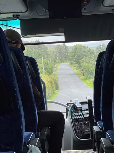 The image provides an interior view from a bus, looking forward through the windshield. The road ahead is wet and appears to be a rural, winding path surrounded by lush green trees and vegetation, with a gray sky suggesting overcast or rainy weather. In the foreground, the dark blue seats of the bus are visible, and a person wearing a baseball cap is seated on the left. The bus's dashboard, with various buttons and a mounted phone, is also visible. The scene is captured from within the bus, implying a journey is in progress. The location is identified as Cearnóg Sheain Bháin, Ireland.
