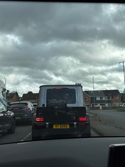 The image captures a ground-level view from inside a vehicle looking forward through the windshield, depicting a traffic scene on a residential/suburban road in Hagley, United Kingdom, under a heavily overcast sky.

In the immediate foreground, the top edge of a vehicle dashboard is visible. Directly ahead, a black and silver (or dark grey) Mercedes-Benz G-Class G 580 SUV is stationary or moving slowly in traffic. Its rear-mounted spare tire cover displays the Mercedes-Benz logo, and both rectangular rear brake lights are illuminated red. The vehicle's yellow rear license plate displays the registration "RC 9999".

Behind the G-Class, slightly to the left, a dark grey SUV with distinct horizontal LED taillights (registration "4 HMD" partially visible on its yellow rear plate) is also stopped in the same lane. Further left, the partial side of a large white and blue truck is visible, along with the rear portion of another dark-colored car in an adjacent lane or further ahead.

The background consists of a row of two-story residential houses lining the right side of the road. These houses feature brick construction, dark pitched roofs, and multiple windows. Beyond the vehicles and houses, a road junction with active traffic lights is visible to the far right. Multiple tall, black streetlights are positioned along the road. The road surface appears to be asphalt with white lane markings. Sparse vegetation, including dry grasses and small trees, is visible along the roadside and between properties. No individuals are discernible in the scene.