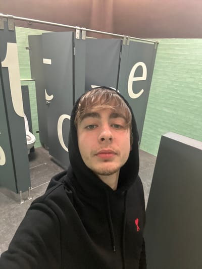 A person with light brown hair and a black hooded sweatshirt is taking a selfie in a bathroom. The person is facing forward with a slightly tilted head. Behind the person are bathroom stalls with large white letters on the gray doors. Light green tiles are visible on the wall above the stalls. The floor is a light gray, possibly tile or concrete. A small red logo is visible on the right chest of the hooded sweatshirt.