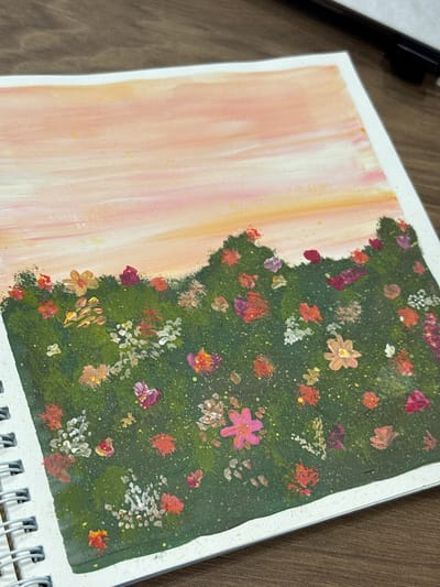 An aerial view of a painting on a wooden surface. The painting depicts a field of flowers against a sunset sky. The sky is rendered in soft strokes of pink, orange, and yellow, with a few scattered yellow dots suggesting starlight or distant light. Below the sky is a dark green, textured band representing a hillside or meadow, adorned with numerous small, impressionistic flowers in shades of red, pink, white, and yellow. The painting is bound in a white spiral notebook, the edge of which is visible on the left side of the frame. The wooden surface appears to be a table or desk. There are no people or animals visible in the image, and no specific location in the city of Murom, Russia, can be identified.