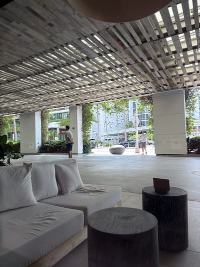 The media file displays an outdoor or semi-outdoor lounging area, likely a common space in a modern building in Miami Beach, United States of America.
**Subjects:** The scene features white outdoor lounge furniture, including a long sofa with multiple cushions, and two large, dark cylindrical tables or stools. On the left, a prominent architectural element consists of a slatted wooden ceiling or wall, possibly a pergola or sun screen, with light visible through its gaps. In the open background, lush tropical foliage, including palm trees and dense bushes, is visible. Two men are present: one is seen walking further into the distance on a paved surface, while another man stands closer to the foreground, facing away from the camera, near some greenery. Modern buildings with large windows are visible in the background. A dark car is parked on a street in the mid-ground.
**Activity or event:** The men appear to be casually traversing or standing in the outdoor space. There is no specific event or interaction clearly depicted. The scene suggests a relaxed atmosphere in a communal area.
**Notable details:** The time of day is bright daylight, indicated by clear lighting and shadows, suggesting a sunny day. The presence of palm trees and tropical greenery is consistent with the Miami Beach location. The architecture appears modern and airy, designed to integrate with the outdoors.
**Visible text:** The word "Avis" is clearly visible in red letters on a white sign in the background, near the standing man.