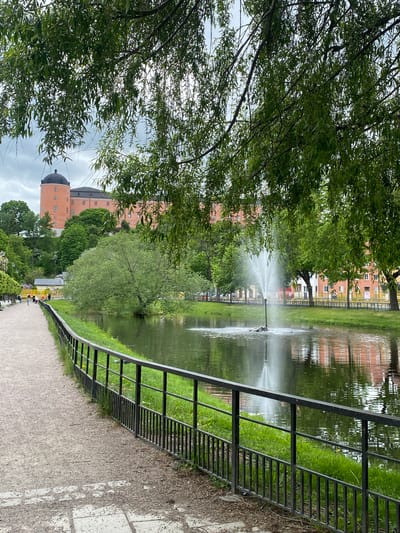 A partly cloudy, daytime outdoor scene depicts a pond and a sidewalk bordering the pond, located in Uppsala, Sweden. In the middle of the pond, a fountain shoots water upwards. The pond reflects buildings and the trees. A black metal fence runs along the sidewalk next to the pond. On the sidewalk, several people are visible in the distance, some sitting on benches. Lush green trees are on the left side of the pond and sidewalk. In the background, an orange-colored castle with a black dome is partially visible through the trees. Further in the background, other buildings of varying colors can be seen. The sidewalk is made of a light-colored material.