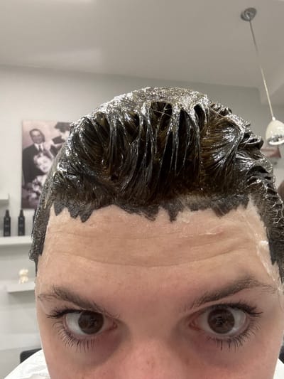 A close-up, upward-angled photograph depicts the face of an individual undergoing a hair treatment within a salon located in Cortona, Italy. The subject's hair is saturated with a thick, dark, viscous substance, consistent with hair dye or a deep conditioning treatment, which extends irregularly onto the forehead and temples. A clear, protective barrier or cream is applied to the skin along the hairline, separating it from the dark product in visible areas. The individual's wide-open, dark-brown eyes gaze directly at the viewer, with reflections consistent with a handheld camera or smartphone. The background features a light-colored wall on the left, displaying a framed black-and-white portrait of multiple individuals, including at least two men in suits and a woman, presented in a vintage style. Below the portrait, a white shelf holds several dark, cylindrical bottles. The ceiling on the right is light-colored, from which a modern, chrome pendant light fixture is suspended.