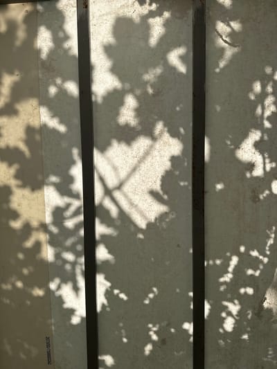 An overhead shot captures a textured, off-white surface, possibly a translucent canopy or tarp, partially obscured by dark shadows cast by foliage and thin, vertical metal beams. The surface appears weathered, with irregular staining and a visible grid-like pattern from its weave. The shadows create an abstract composition, with dappled light filtering through in places. A faint series of numbers is printed in the lower left corner. The overall impression is one of an outdoor setting, likely under a structure providing partial shade, such as a pergola or a covered seating area in Завалочыцкі сельскі Савет, Belarus.