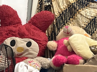 The image shows a close-up of several stuffed toys, appearing to be in a domestic setting. The stuffed toys are predominantly red and pink, with some beige accents. One large red stuffed toy with large black eyes and a yellow nose, resembling a character from a children's show, is positioned on the left side of the frame. To its right, a pink stuffed animal with beige ears and paws is visible. Behind the toys, a patterned fabric with dark blue and gold designs is draped. In the foreground, a black fan grille is partially visible on the left, and a white piece of fabric is draped over something on the bottom left. A cardboard box is also visible at the bottom right of the image, containing some of the stuffed toys. The lighting suggests an indoor environment, possibly a bedroom or playroom. The location in Cadek, Indonesia is not discernible from the visual information in the image.