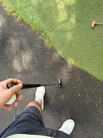 A person in white sneakers and gray shorts stands on a dark paved surface speckled with small debris, holding a golf ball in one hand and a putter in the other. A green artificial turf section is adjacent to the paved area, with an orange golf putter lying on it. The scene appears to be a miniature golf course, possibly located in Warrington, United Kingdom, though definitive city identification is impossible from the image alone.