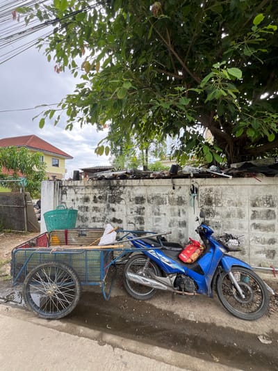 In the city of Ban Kanchanok 23, Thailand, a blue motorcycle is parked next to a two-wheeled cart. The cart is filled with items, including a green basket, bamboo poles, and a white bag. The motorcycle has a basket on the front containing a red bag of goods. Behind the cart and motorcycle is a rough, white cinder block wall, topped with debris and vegetation. A large tree with dense foliage overhangs the scene from the right. In the background, a yellow house with a red roof is visible, along with a chain-link fence. The ground is a mix of dirt, concrete, and puddles of water, suggesting recent rain. Overhead, electrical wires are strung between poles. The overall scene depicts a cluttered, outdoor environment, possibly a residential or commercial area.