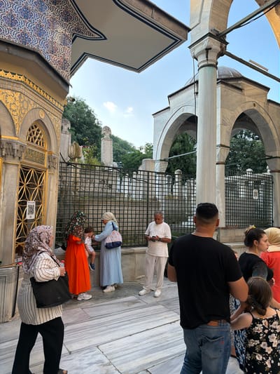 The image depicts an exterior scene at the Eyüpsultan Mosque complex in Eyüpsultan, Turkey. Several individuals are present, engaged in various activities.
In the foreground, a person wearing a black t-shirt and blue jeans is seen from behind, facing away from the camera. To their right, a woman in a black top with a floral pattern, partially visible, stands alongside another woman in an orange dress, who is also largely out of frame. A young child is seated on a stone ledge.
Further to the left, a woman in a striped long-sleeved shirt and black pants, carrying a black tote bag, walks away from the camera. Next to her, a woman in a vibrant orange dress and a headscarf is interacting with the child. Another woman in a light blue long dress and headscarf stands near the child, holding a bag with blue and white stripes. To their left, a man in a white shirt and pants is looking at his phone.
The background reveals architectural elements characteristic of the mosque complex, including arched structures, marble columns, and ornate tiling on a building facade. Beyond the architectural features, a black metal fence encloses a graveyard with numerous gravestones and trees. The sky above is clear with a few wispy clouds. The ground is paved with large marble slabs. The overall atmosphere suggests a visit to a significant religious and historical site.