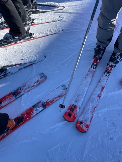 The image provides an overhead view of skiers' legs and equipment on a bright, snow-covered ski slope in Séez, France. At least three individuals are depicted through their ski gear. In the upper part of the frame, a person wearing grey ski pants and black ski boots is standing with their red and black skis, which are partially covered in powdery snow. A silver ski pole extends diagonally from this skier towards the lower right.
Below, multiple pairs of skis are visible resting in the snow. One pair of red and black skis, with "ARMADA" clearly visible on its surface, is prominent in the mid-ground. Another pair of red, black, and white skis, showing fragmented text like "BLITRA" and "900S", is positioned towards the lower right, connected to orange and black ski boots. A third set of black-clad legs and skis can be seen in the lower-left corner.
The scene suggests a moment of rest or preparation during a ski outing. The snow is bright white and textured, with some areas appearing freshly fallen or undisturbed, while others show tracks and compacted surfaces. The strong shadows cast by the equipment indicate bright daylight and clear weather. The skis and boots are dusted with snow, highlighting the cold, wintry conditions of the mountain environment.