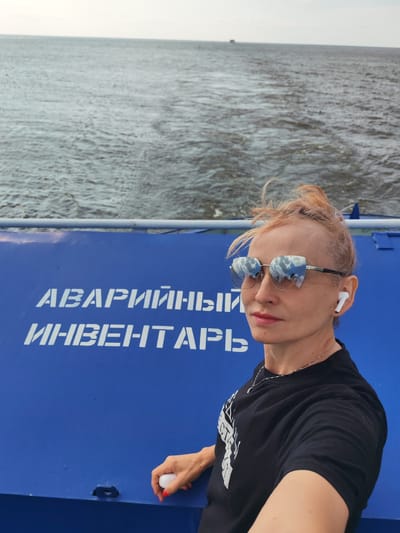 A medium shot shows a blonde woman with her hair pulled back in a bun, wearing sunglasses, a black t-shirt, and a necklace. She has white wireless earbuds in her ears and is holding a small white object in her right hand. She is standing on a blue surface that appears to be part of a ship or boat, with the word "АВАРИЙНЫЙ ИНВЕНТАРЬ" (Emergency Inventory) written in white letters. The background is a body of water, likely a river or lake, with a faint horizon line and a distant boat. The image was taken outdoors, possibly in the city of Krokhino, Russia, given the context of the Russian text on the surface.