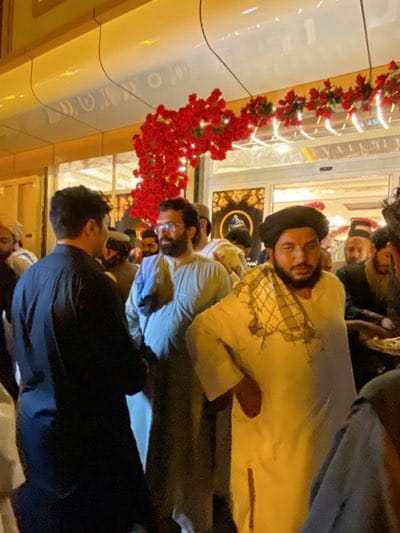 Here is a detailed description of the image:
**Scene:** A group of men stand in front of a building decorated with red floral arrangements above the entrance.
**People:** The men are dressed in a mix of traditional and casual attire. Some wear head coverings, including turbans and kufis. Several have beards. They are standing and facing different directions, suggesting a casual gathering or conversation.
**Location:** The city is likely قندهار, Afghanistan, based on the clothing and appearance of the men.
**Objects:** The building in the background appears to be a commercial establishment. The red floral arrangements add a celebratory or decorative element.
**Action/Interaction:** The men appear to be conversing and milling around. The overall scene gives the impression of a social gathering.
**Environment:** The lighting suggests that the photograph was taken in the evening or under artificial light. The environment seems to be an urban setting.