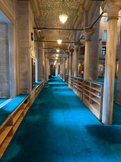 This image depicts the interior of a mosque, likely in Eyüpsultan, Turkey, given the architectural style. A long hallway is lined with massive stone columns on the right, supporting an ornately decorated ceiling with intricate patterns and dangling pendant lights. On the left, the hallway features a stone wall with an archway and windows. Along the base of the wall and extending down the hallway, there are wooden shelves built into benches, covered with bright turquoise cushions. The floor is covered with a continuous, dark turquoise carpet. Several pairs of shoes are visible on the shelves. In the distance, at the end of the hallway, a person is seated, reading. The lighting is dim, with light emanating from the pendant lamps. The overall impression is one of solemnity and architectural grandeur.