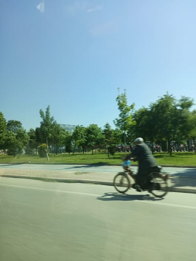 A person is riding a bicycle on a paved path beside a grassy area with trees. The person is wearing a dark jacket and a light-colored cap. In the background, there are more trees, and a building with a skeletal structure is visible. Several red tractors are parked in the distance to the left. The sky is clear and blue. The image appears to have been taken from a moving vehicle, as indicated by the motion blur. The scene is likely in Karatay, Turkey, given the context provided.