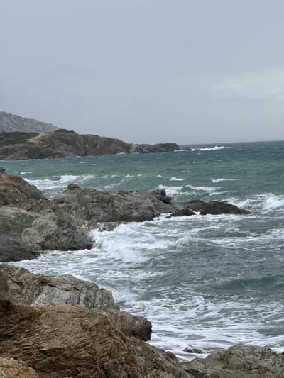 The image depicts a turbulent coastal scene within the Saronida Municipal Unit, Greece, under an overcast sky. In the foreground, rugged, stratified grey and reddish-brown rock formations dominate the lower left and center, extending into the midground. The sea, colored in shades of dark teal and grey-green, displays numerous white-capped waves actively breaking against these rocky outcrops, generating significant foam and spray. The wave action suggests rough sea conditions. Towards the left midground, the coastline extends into a series of darker grey rocky headlands and hills, with sparse, muted green vegetation visible on the upper slopes. A faint, lighter-colored track or path is discernable on one of the distant hills. The sky above is uniformly light grey, indicating heavy cloud cover. No individuals or man-made structures are visible.